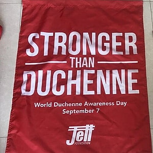 Red house flag with white text that says Stronger than Duchenne at the top in large text.  Below in smaller white text it says World Duchenne Awareness Day September 7.  The Jett logo is below all the text and at the very bottom of the flag in red text with a white background is the website address