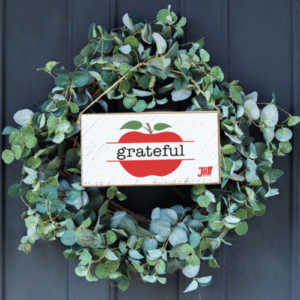 A wreath with a white sign on it. The sign has a red apple with the word grateful in the middle of the apple
